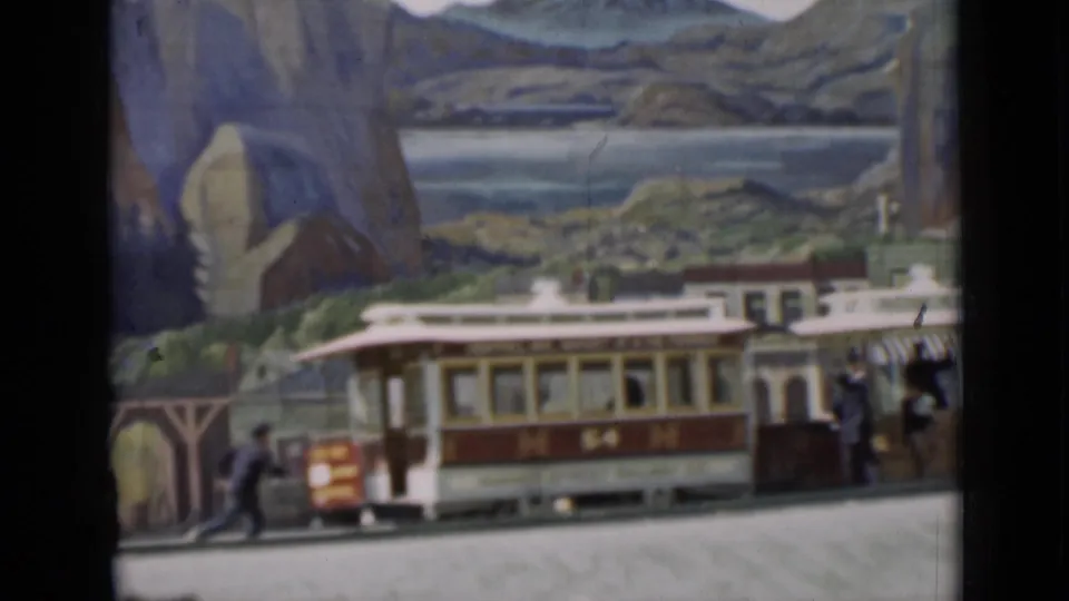 1937 8mm frame of a staged miniature town with a trolley marked M 54, tiny figures, storefronts, and painted mountains behind it.