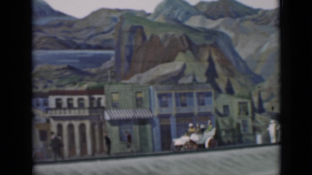 Staged miniature town scene with storefronts, striped awnings, tiny figures, a small vehicle, and painted mountains and water in the background.