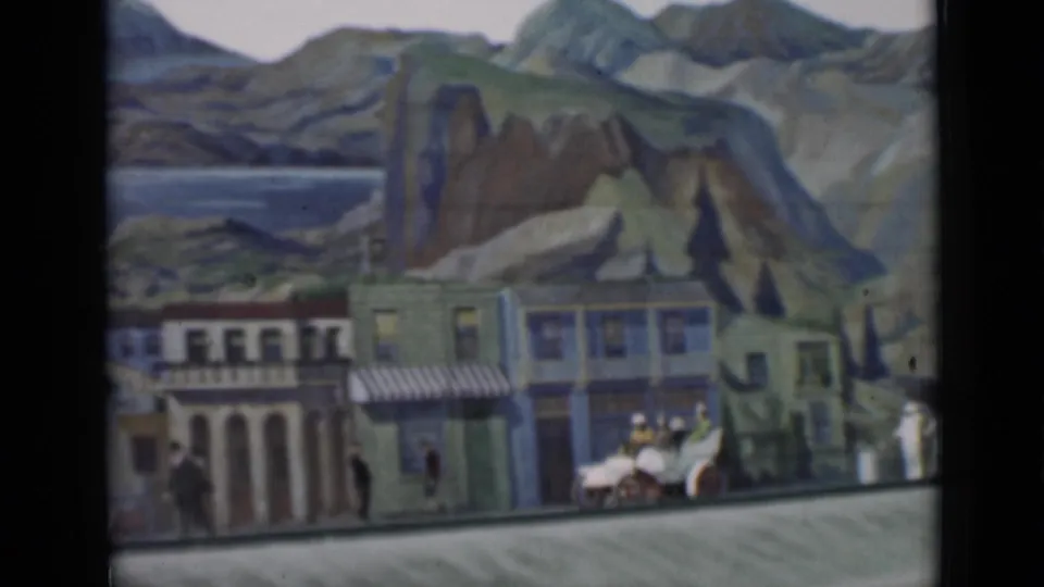 Staged miniature town scene with storefronts, striped awnings, tiny figures, a small vehicle, and painted mountains and water in the background.