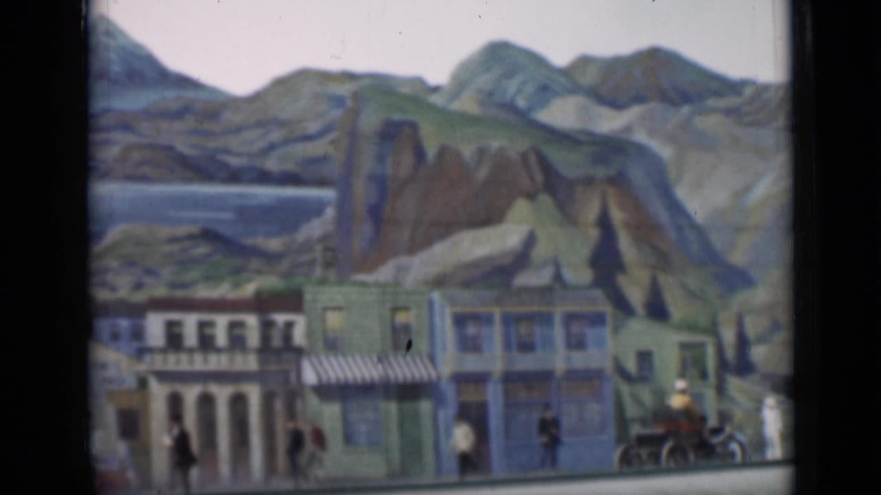 Final 1937 8mm frame of the miniature street display, showing storefront facades, tiny figures, a small vehicle, and painted mountain scenery.