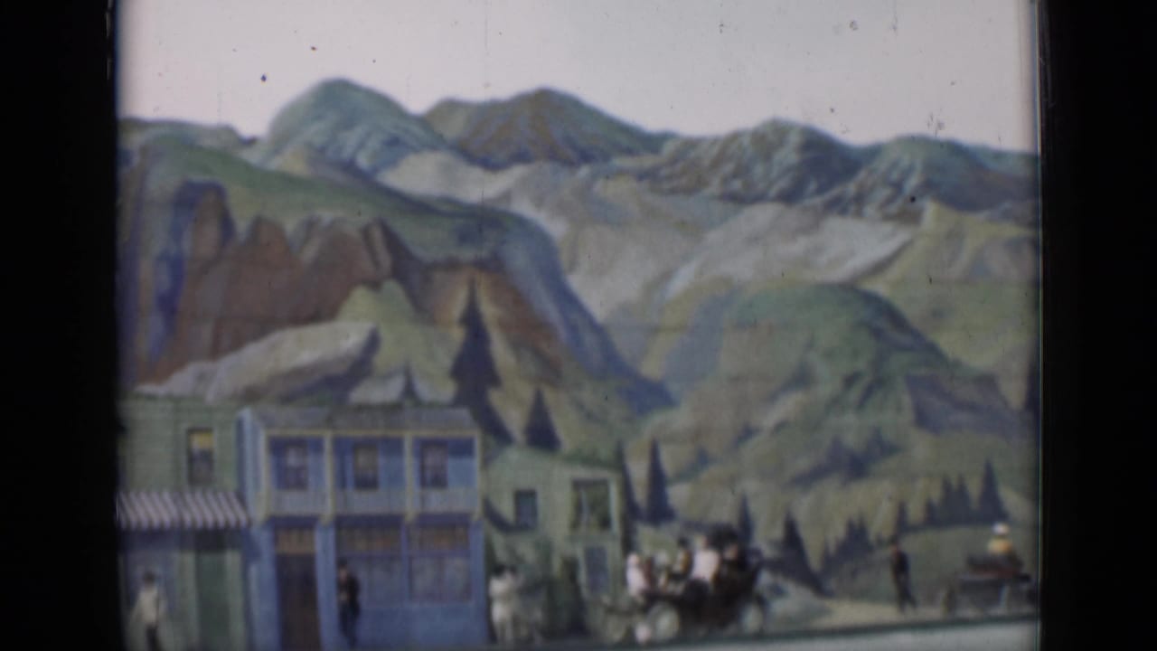 Town facades and painted mountains with pedestrians, a carriage, and a small vehicle at the right.
