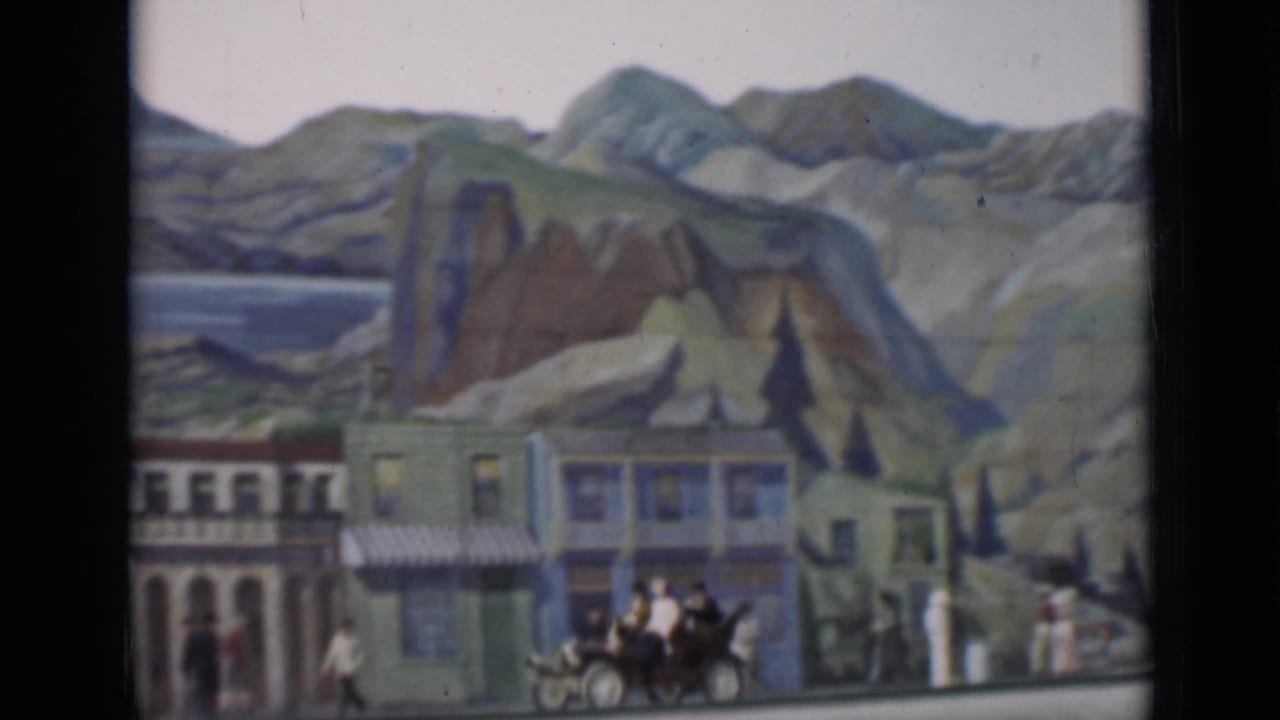 1937 San Francisco 8mm frame showing storefront facades before a painted landscape, with pedestrians, carriage traffic, and an early car.