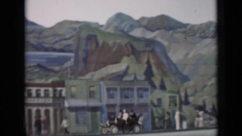 1937 San Francisco 8mm frame showing storefront facades before a painted landscape, with pedestrians, carriage traffic, and an early car.