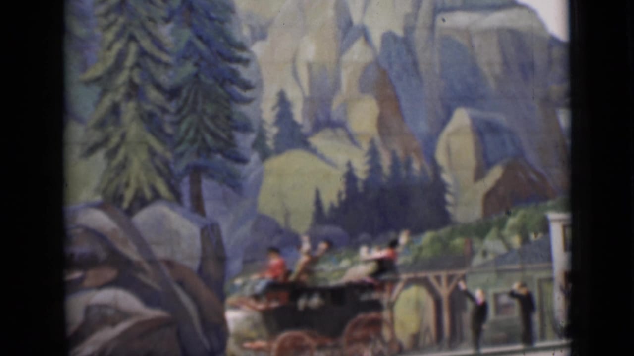 A horse-drawn carriage with riders crosses a studio-style scenic set with large painted rocks, trees, and a mountainous background.