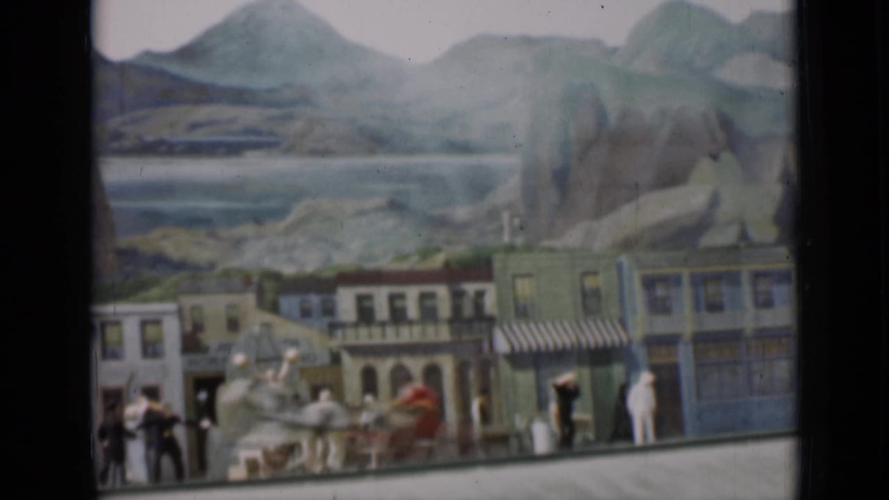 Frame from the 1937 8mm clip showing storefront facades, small figures, horses, and a painted mountain landscape behind the set.