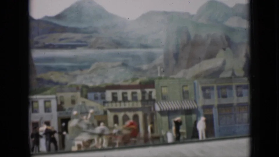 Frame from the 1937 8mm clip showing storefront facades, small figures, horses, and a painted mountain landscape behind the set.