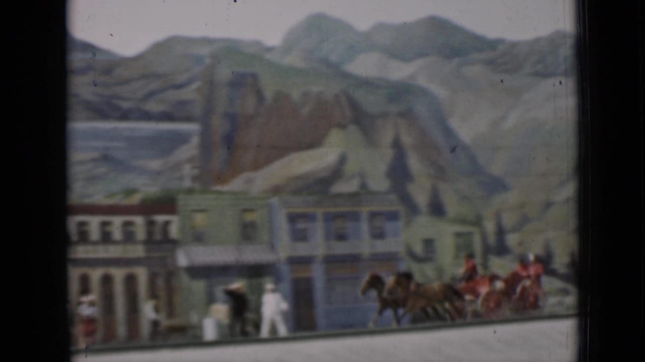 The carriage team appears near the edge of a staged town set, with riders, bystanders, colorful facades, and painted mountains visible.