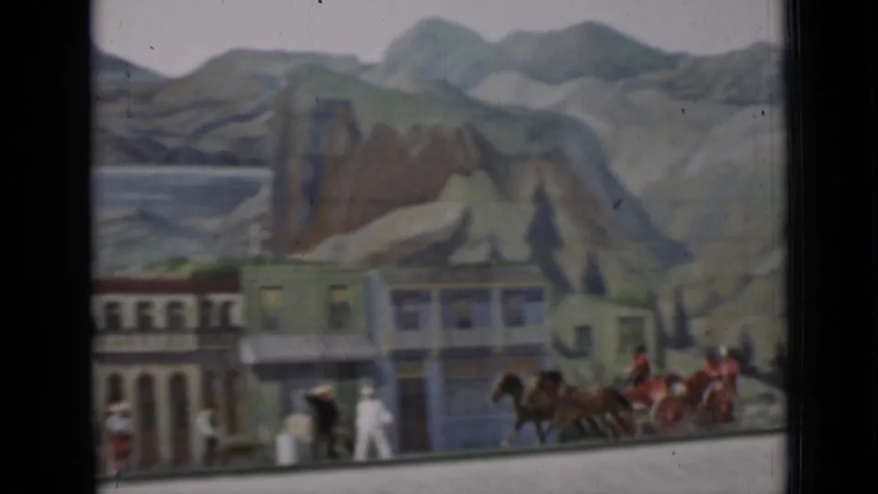 The carriage team appears near the edge of a staged town set, with riders, bystanders, colorful facades, and painted mountains visible.