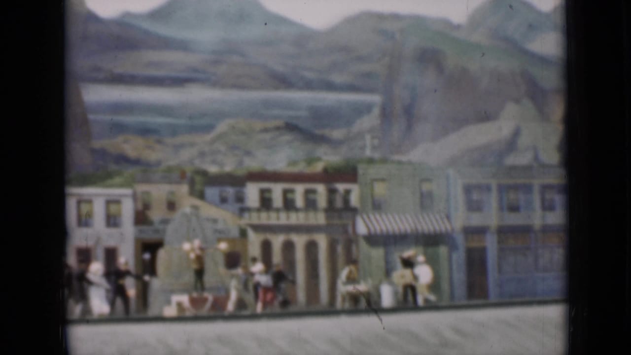 1937 San Francisco 8mm frame of a miniature Western town facade with tiny figures, horses, storefronts, and scenic painted mountains.