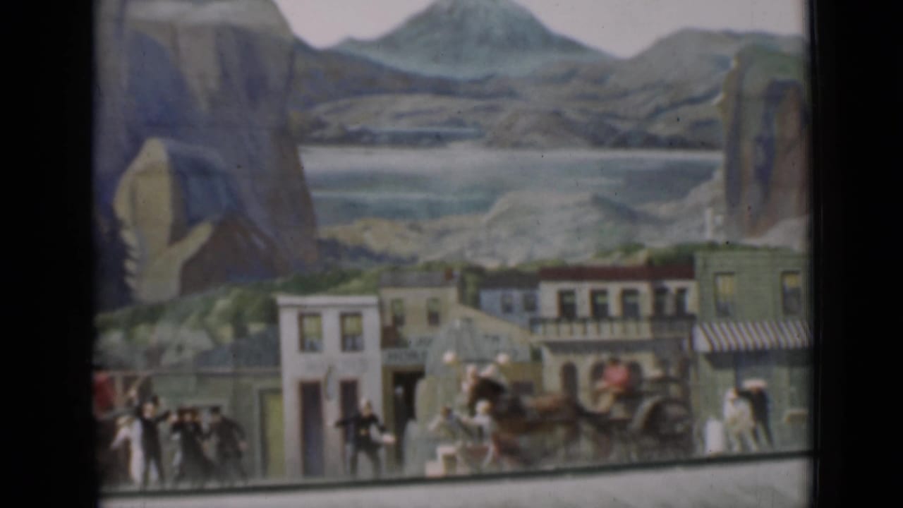 1937 San Francisco 8mm frame of a miniature Western street with a covered wagon or carriage, small figures, storefronts, and painted scenery.