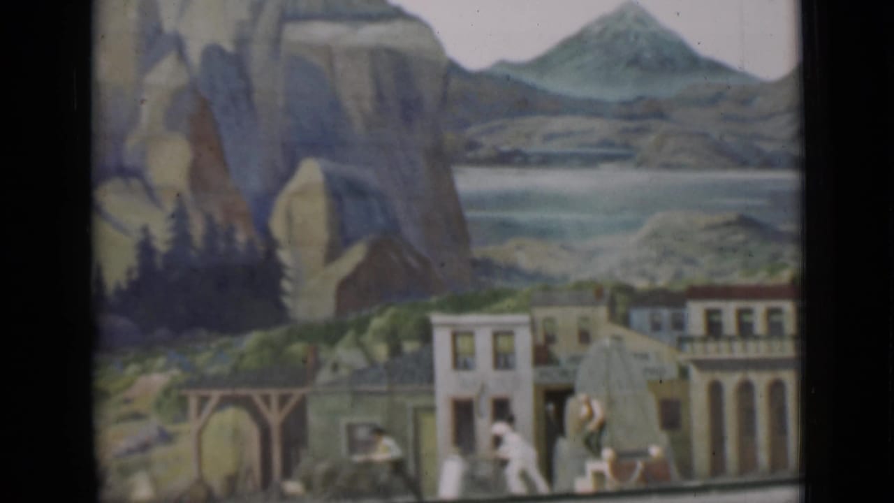 1937 San Francisco 8mm frame of a staged miniature town with painted mountain scenery, small buildings, figures, and a bridge-like arch.