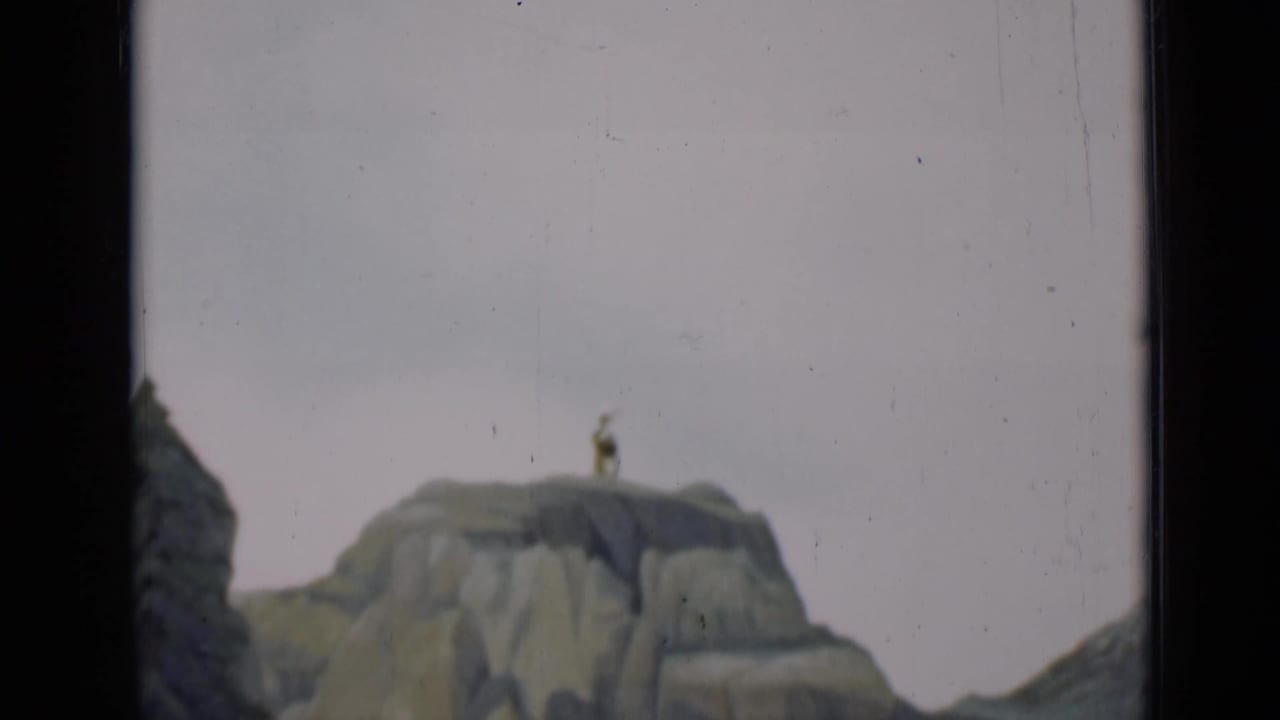 1937 San Francisco 8mm frame showing a rocky scenic model with a small figure posed on a high cliff against a pale backdrop.