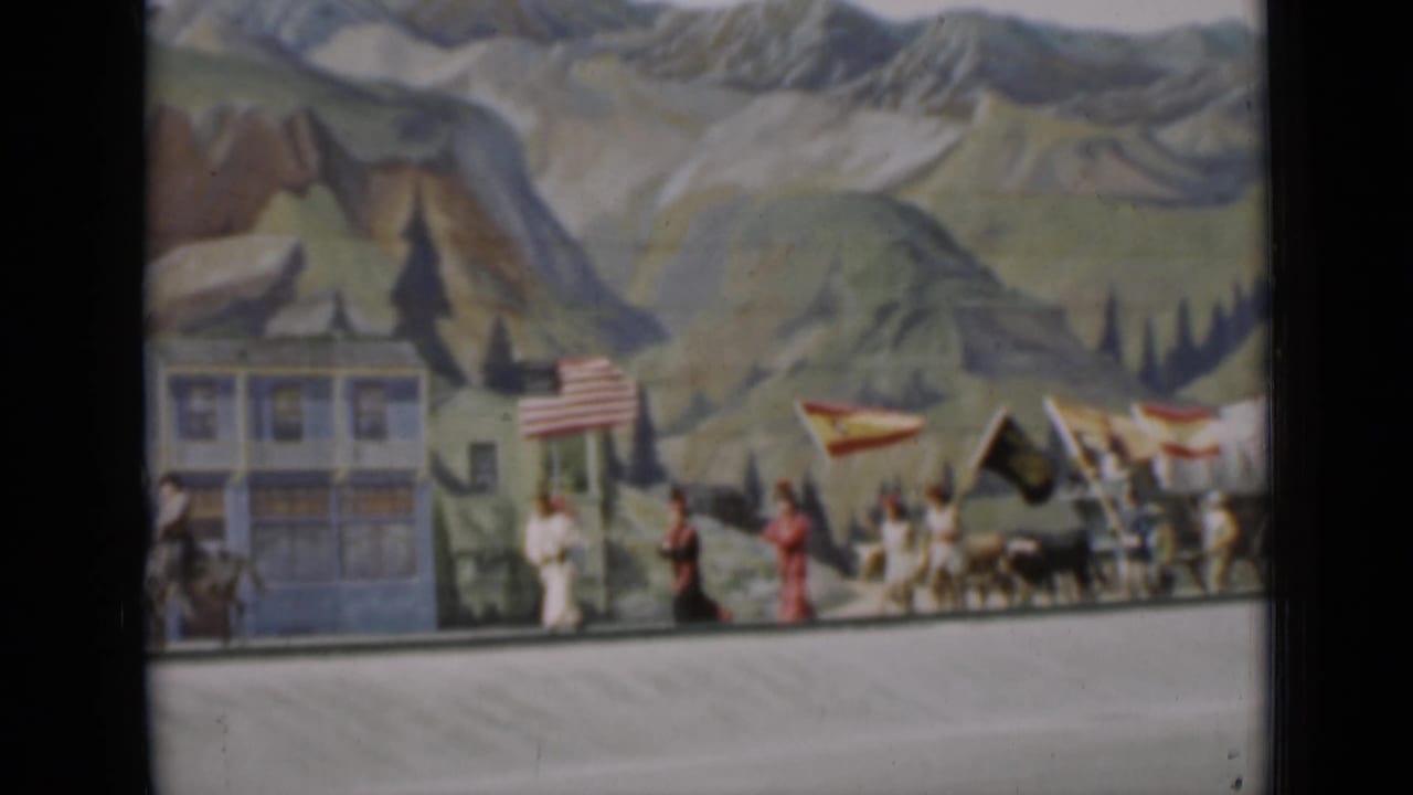1937 San Francisco 8mm frame showing a small flag-bearing procession with figures, animals, and painted scenery.