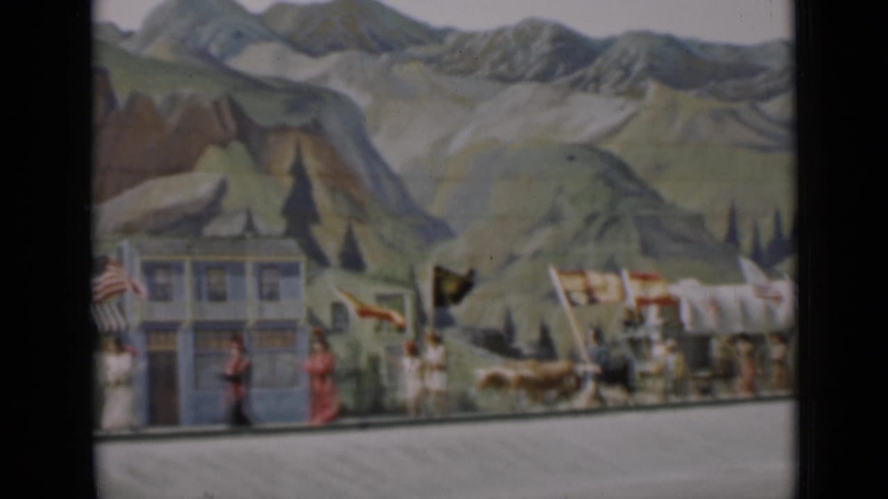 Parade participants carry flags beside a building and painted mountain backdrop.