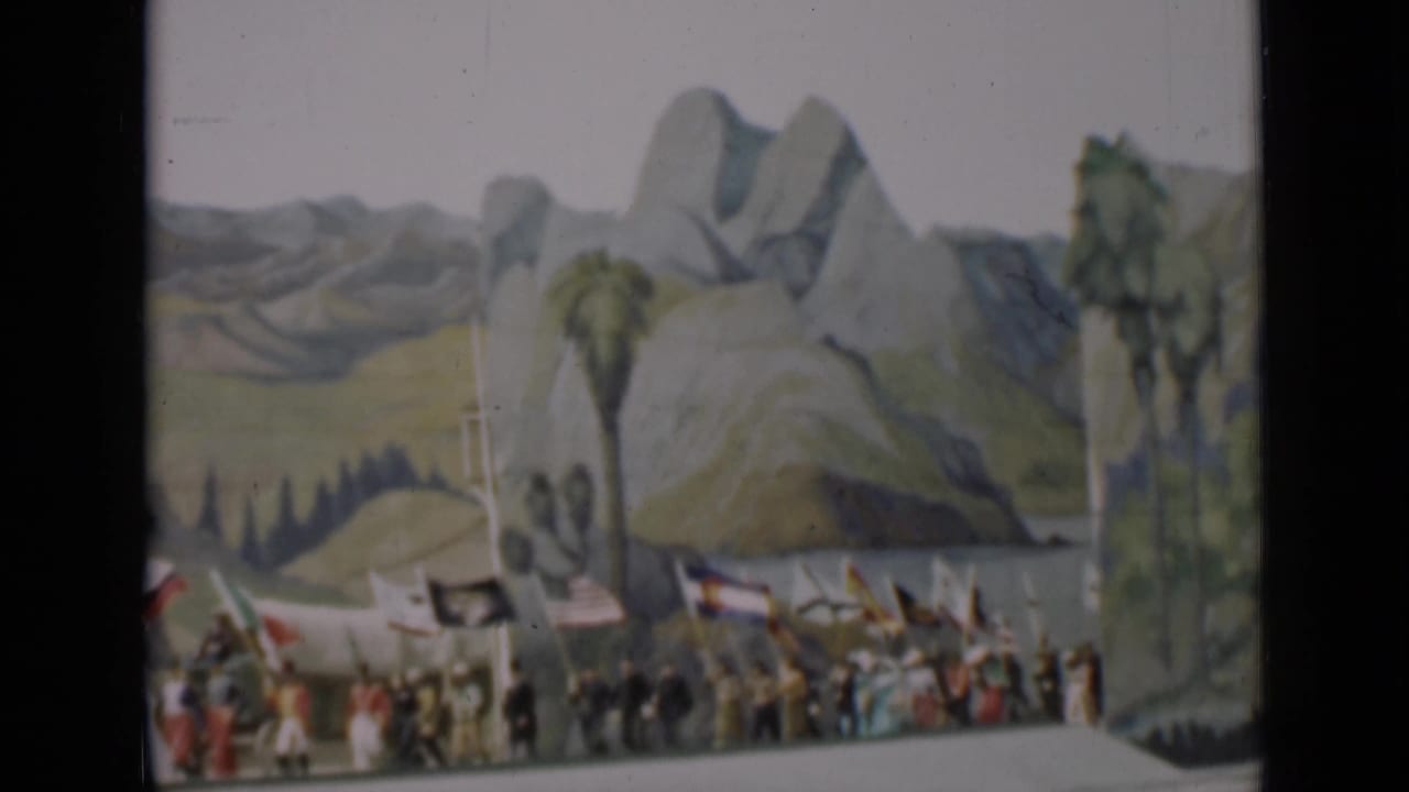 A line of participants carries flags across a stage with mountains painted behind them.