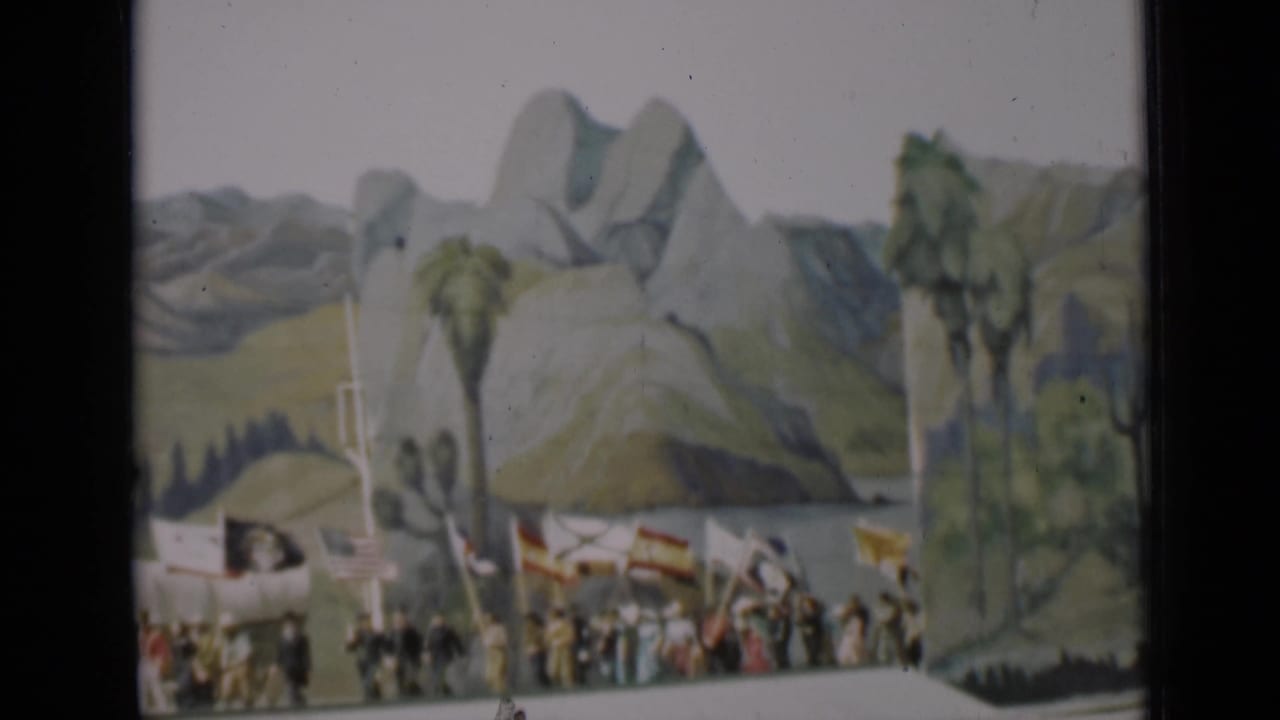 Home-movie frame of a 1937 San Francisco flag pageant, with participants partly blurred as they pass a painted outdoor scene.