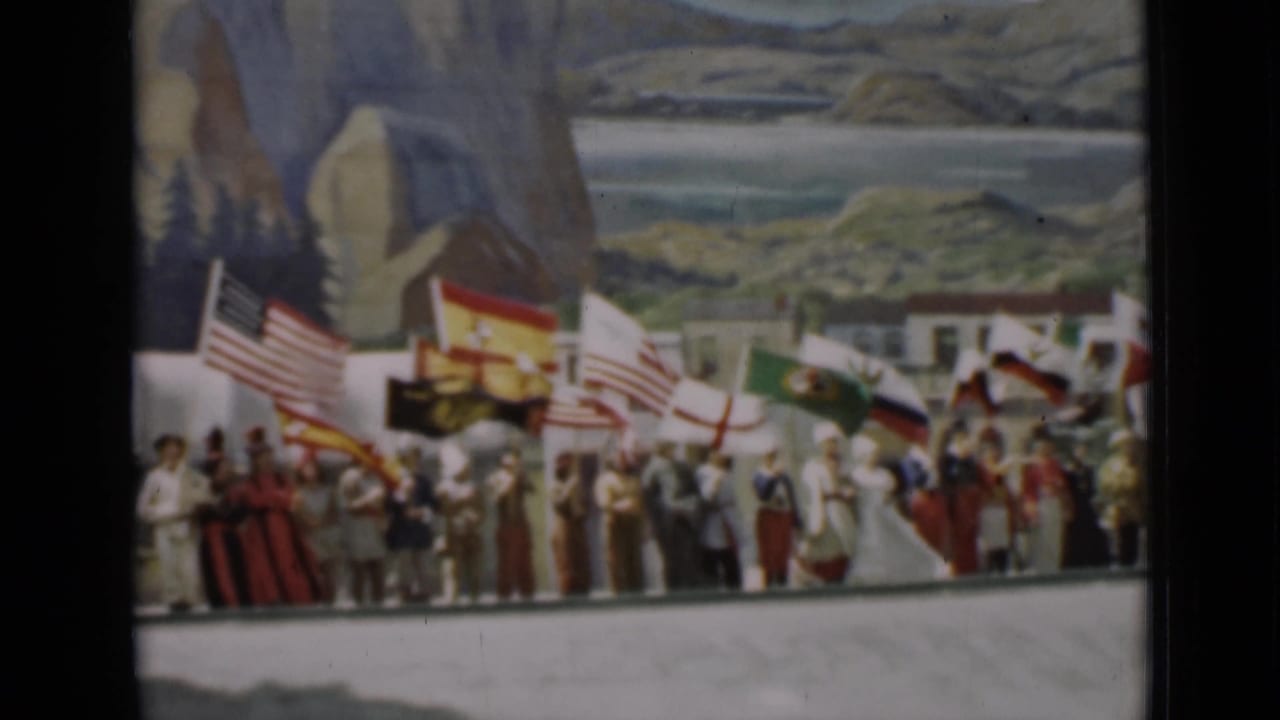 People stand in formation holding national flags before a painted coastal mountain backdrop.