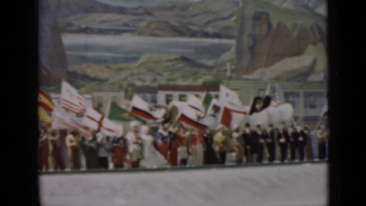 1937 San Francisco frame with national flags, formally dressed participants, and uniformed men arranged before a painted mountain-and-coast scene.