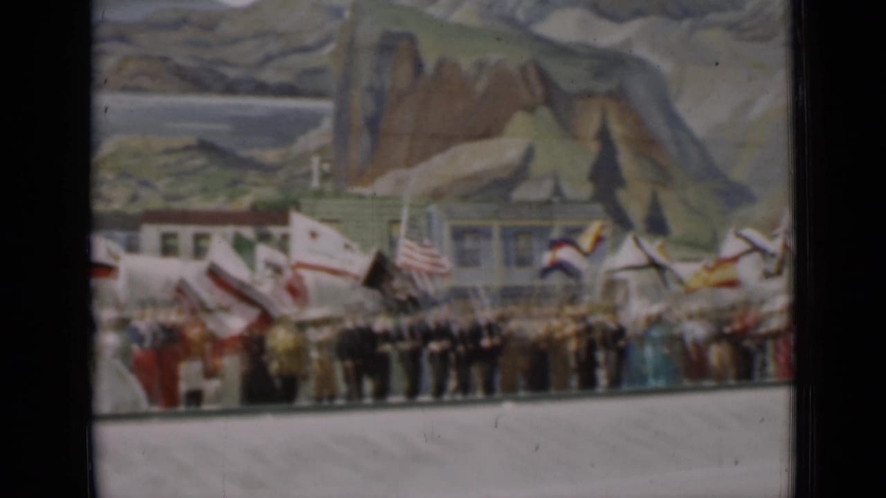 1937 San Francisco home-movie still showing a row of participants and flags beneath a large painted scenic backdrop.
