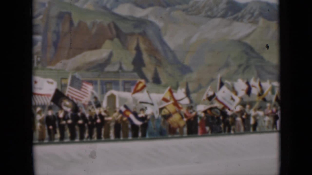 1937 San Francisco frame showing flag-bearing participants, possible uniformed men, and a painted mountain landscape backdrop at a public event.