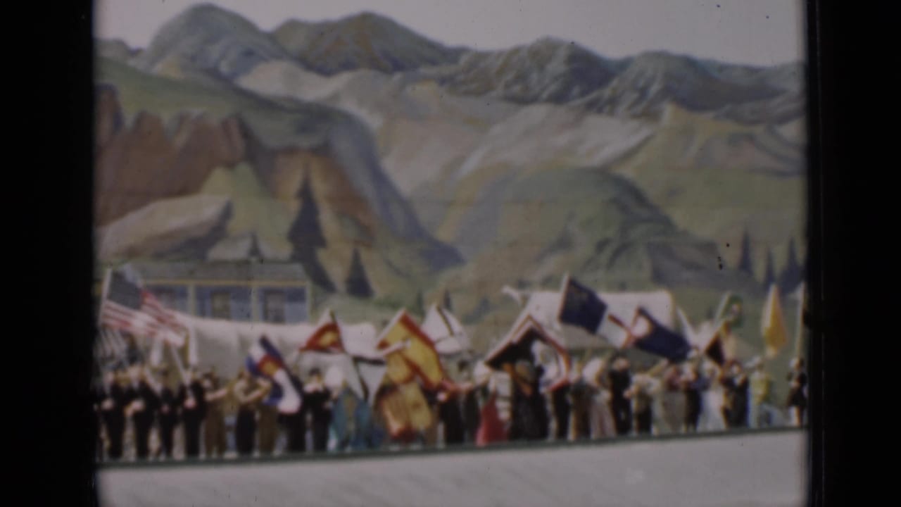 Crowd waving colorful flags before a painted mountain backdrop.