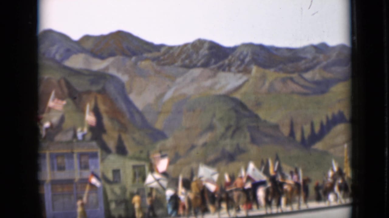 Riders and marching figures pass in front of Western-style buildings and a painted mountain backdrop.