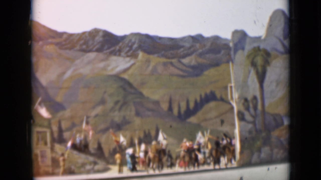 1937 San Francisco 8mm frame showing the procession near a road, with riders, flags, buildings, and painted scenery visible.