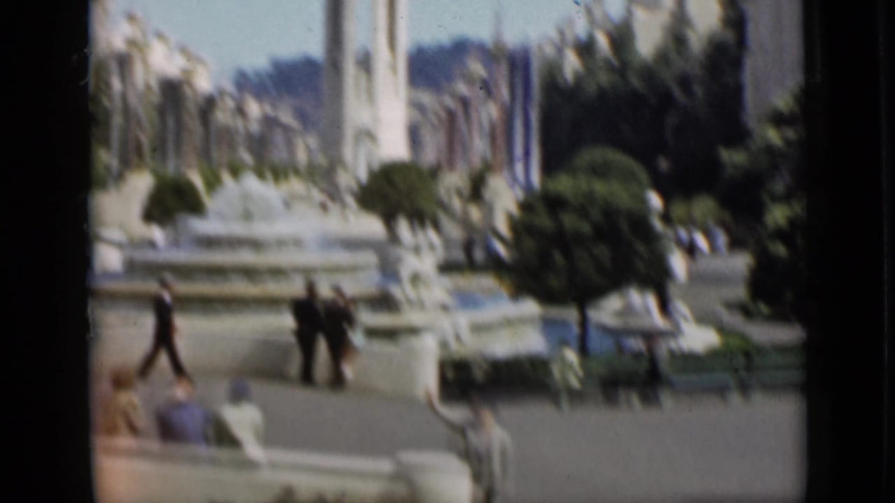Soft 8mm frame of a San Francisco garden plaza with people walking near fountain basins, benches, clipped trees, and a tall pale vertical feature.