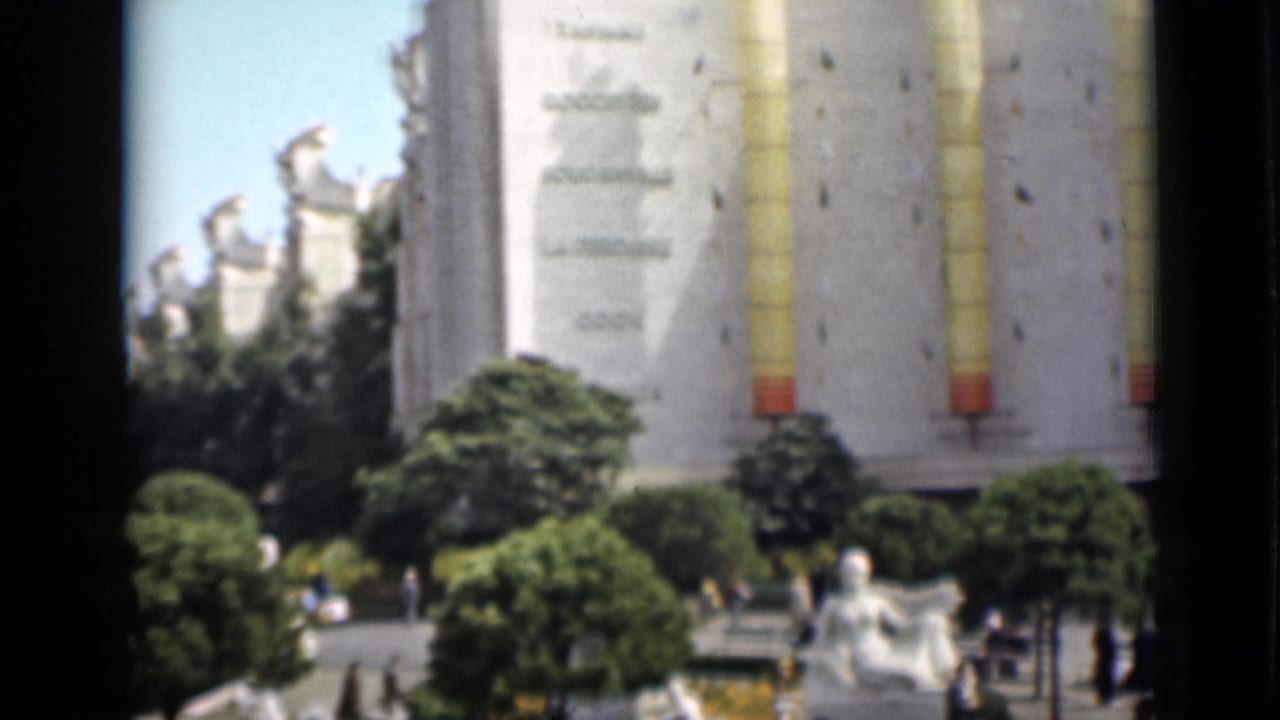 Tower and fairground buildings behind trees, statues, and a plaza fountain.