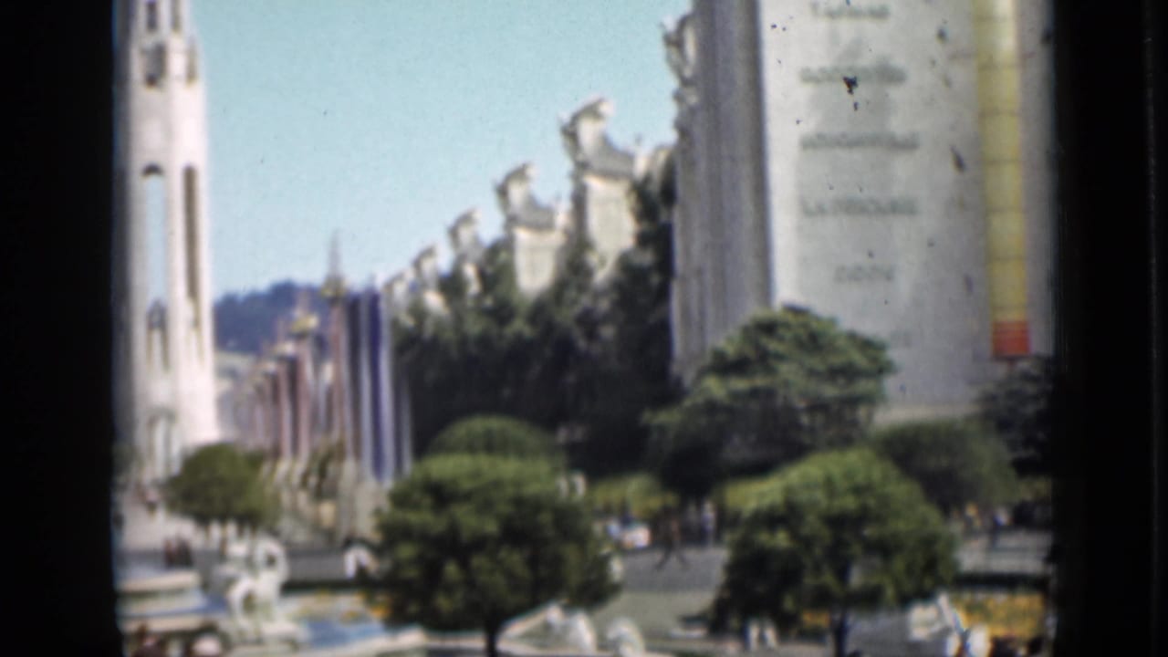 Tall tower, fair buildings, trees, flags, fountain area, and visitors.