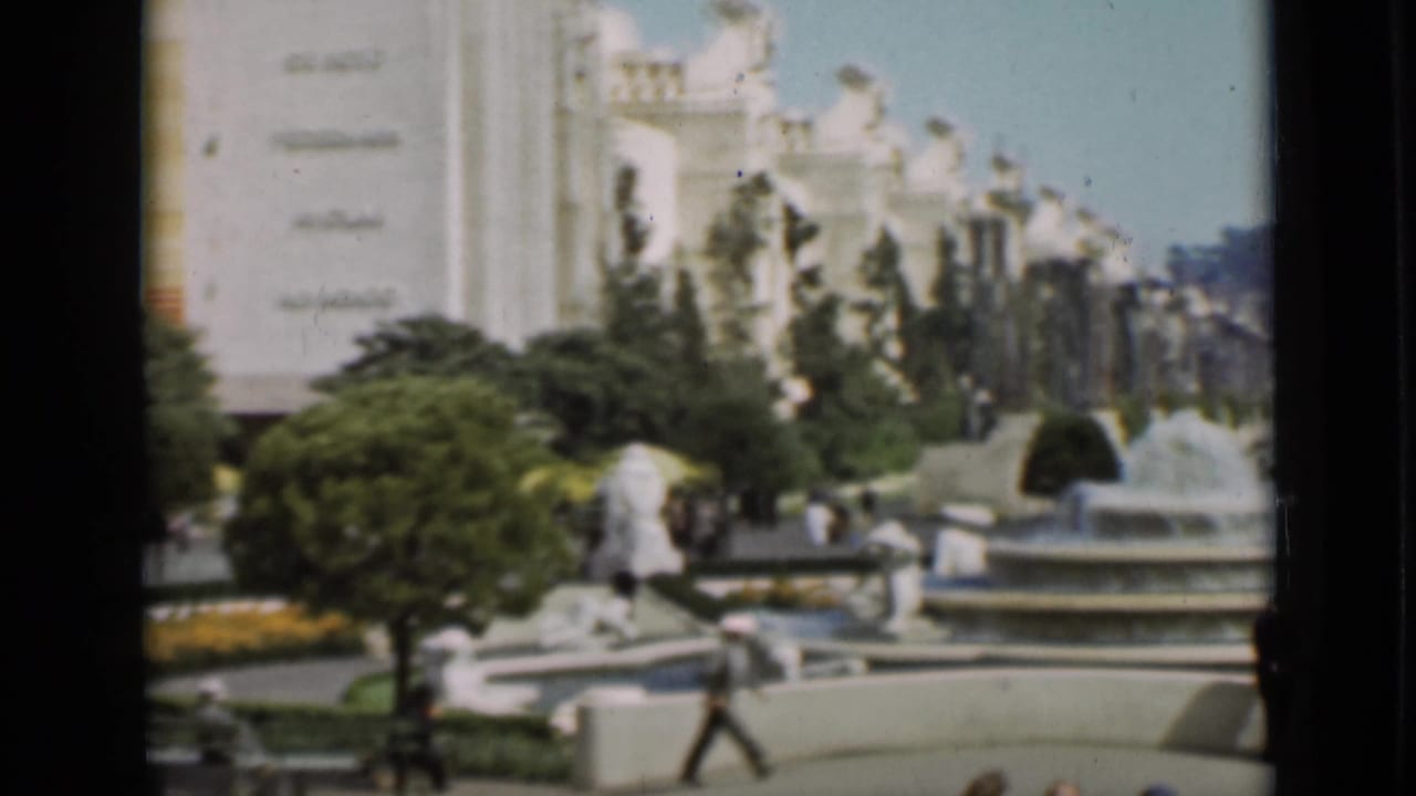 Wide view of a plaza with white buildings, fountains, gardens, and pedestrians.