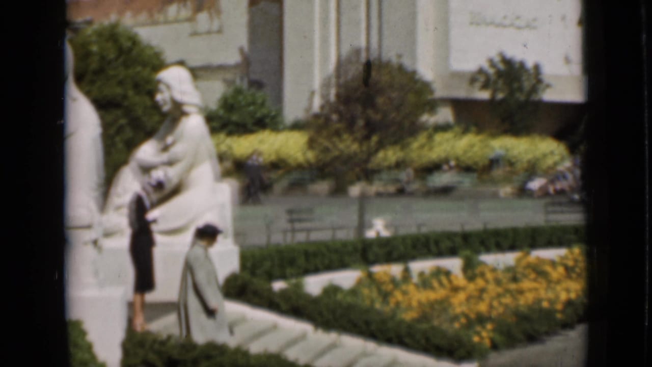 Visitors in period clothing pass white statuary and manicured beds in a San Francisco public plaza, filmed on 8mm in 1937.
