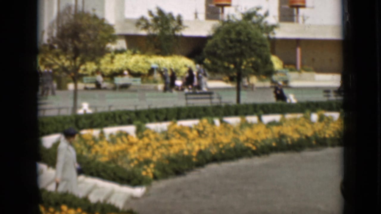 A San Francisco plaza garden scene shows yellow flower beds, benches, hedges, and pedestrians moving through the civic landscape.