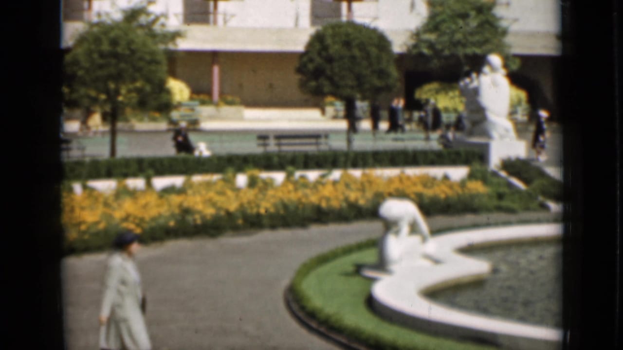 Woman in a long coat walks near a formal fountain with white statues and yellow flowerbeds.