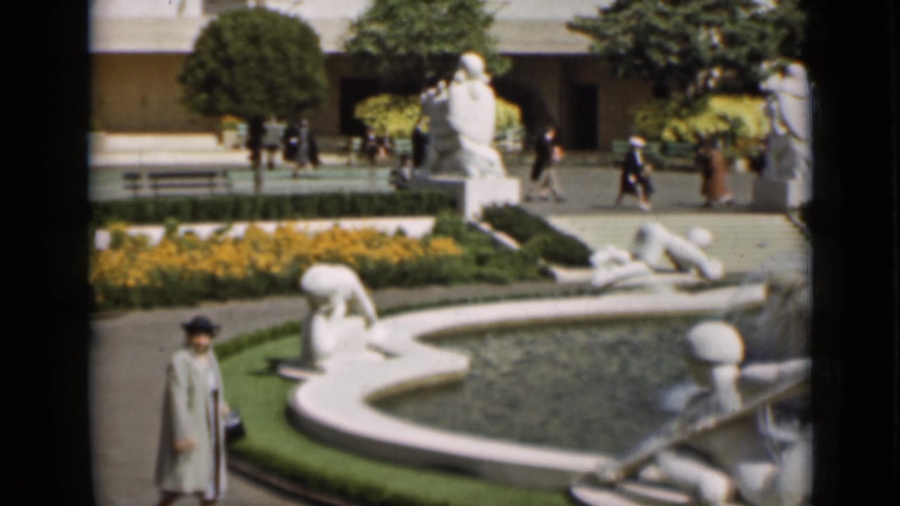 Woman approaches the camera beside a fountain with white sculptures and water behind her.
