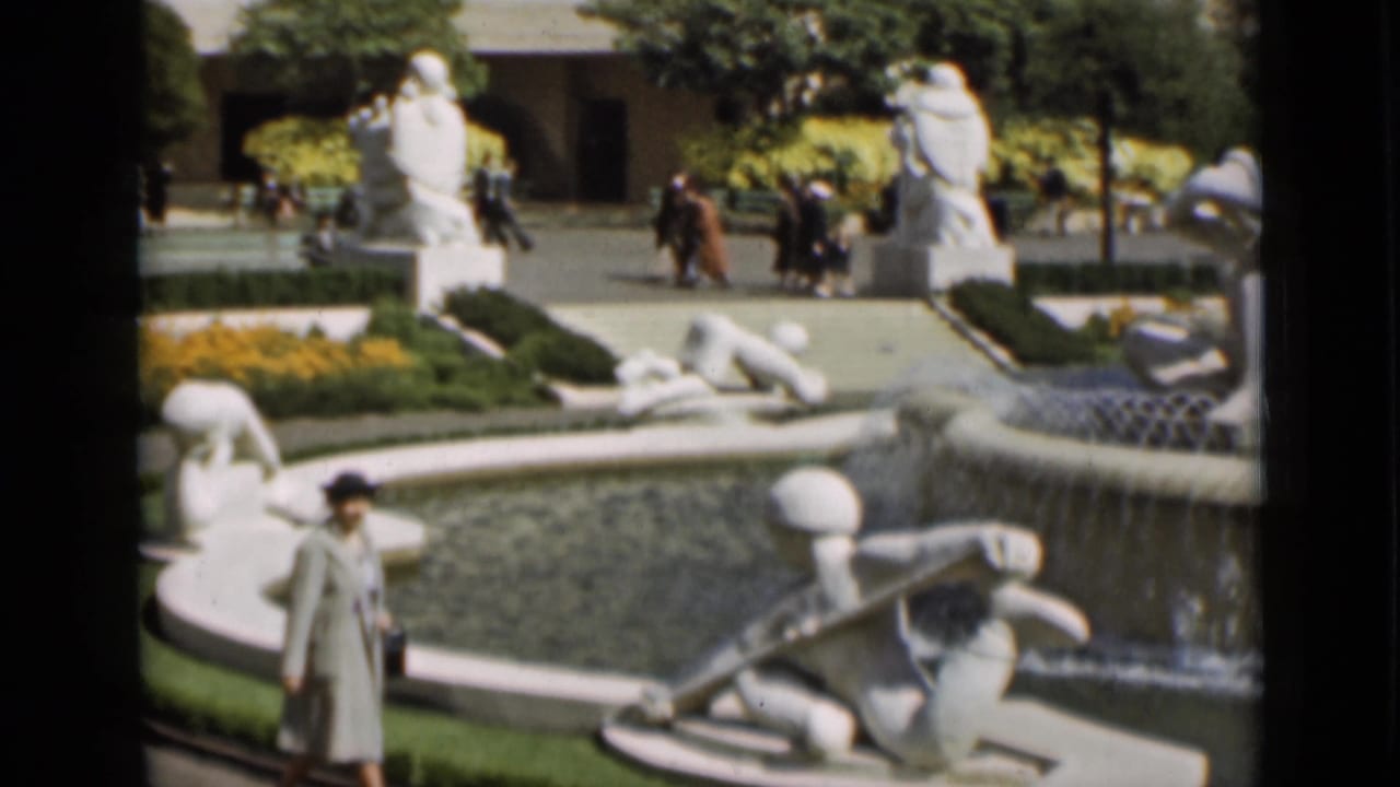 1937 San Francisco home-movie still showing the woman near the foreground of a formal fountain with white statues and visitors beyond.