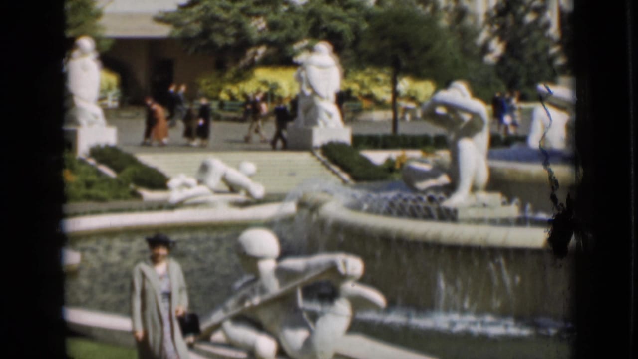 1937 San Francisco frame showing the woman near a fountain with white sculptural figures, flowing water, trees, and visitors in the background.