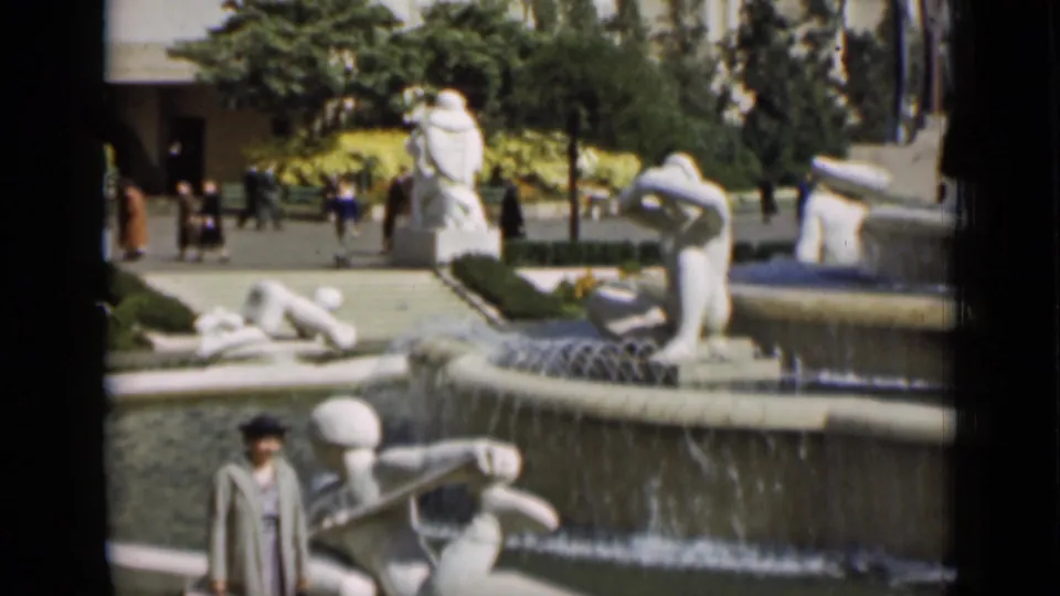 1937 San Francisco home-movie frame showing a tiered public fountain, white sculpture, trees, and a visitor in period clothing.