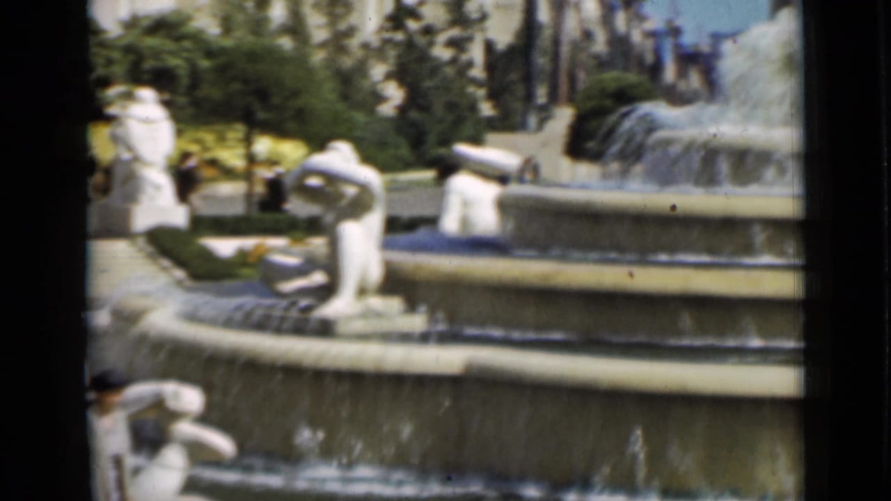 Home-movie view of a San Francisco fountain with white sculptural figures, falling water, trees, and building edges in the background.