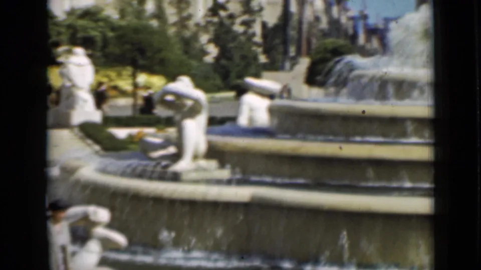 Home-movie view of a San Francisco fountain with white sculptural figures, falling water, trees, and building edges in the background.