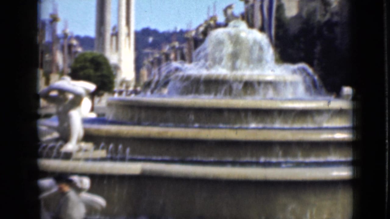 Home-movie frame of the San Francisco fountain from a side angle, showing cascading water, sculpture, trees, and distant hillside.