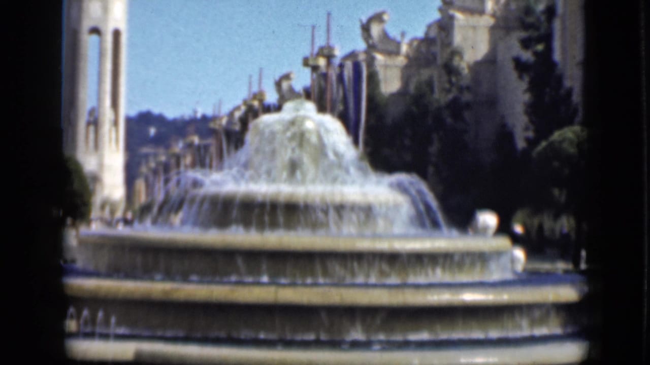 Tiered fountain with water spray and a tall white spire behind it.