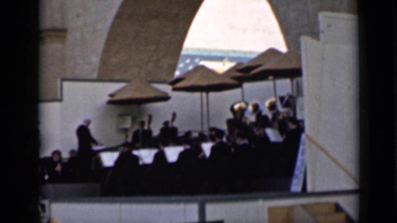 1930s San Francisco home-movie frame showing an outdoor orchestra on a stage, led by a conductor beneath large umbrellas.