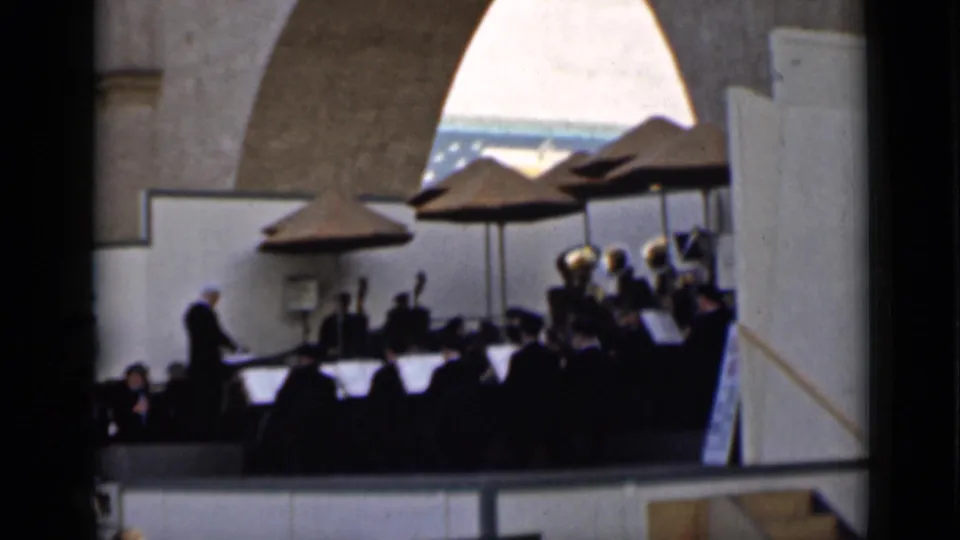 1930s San Francisco home-movie frame showing an outdoor orchestra on a stage, led by a conductor beneath large umbrellas.