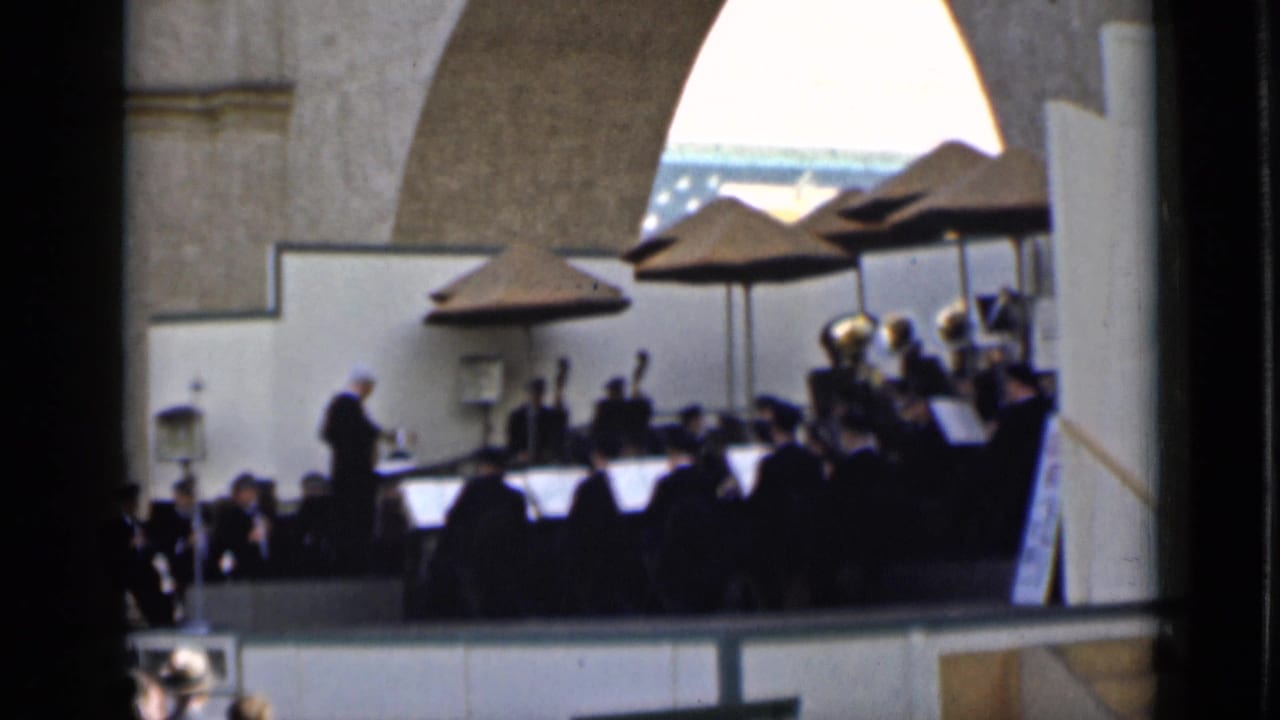 Conductor leads a seated band on a raised outdoor stage beneath umbrellas and stone arches.