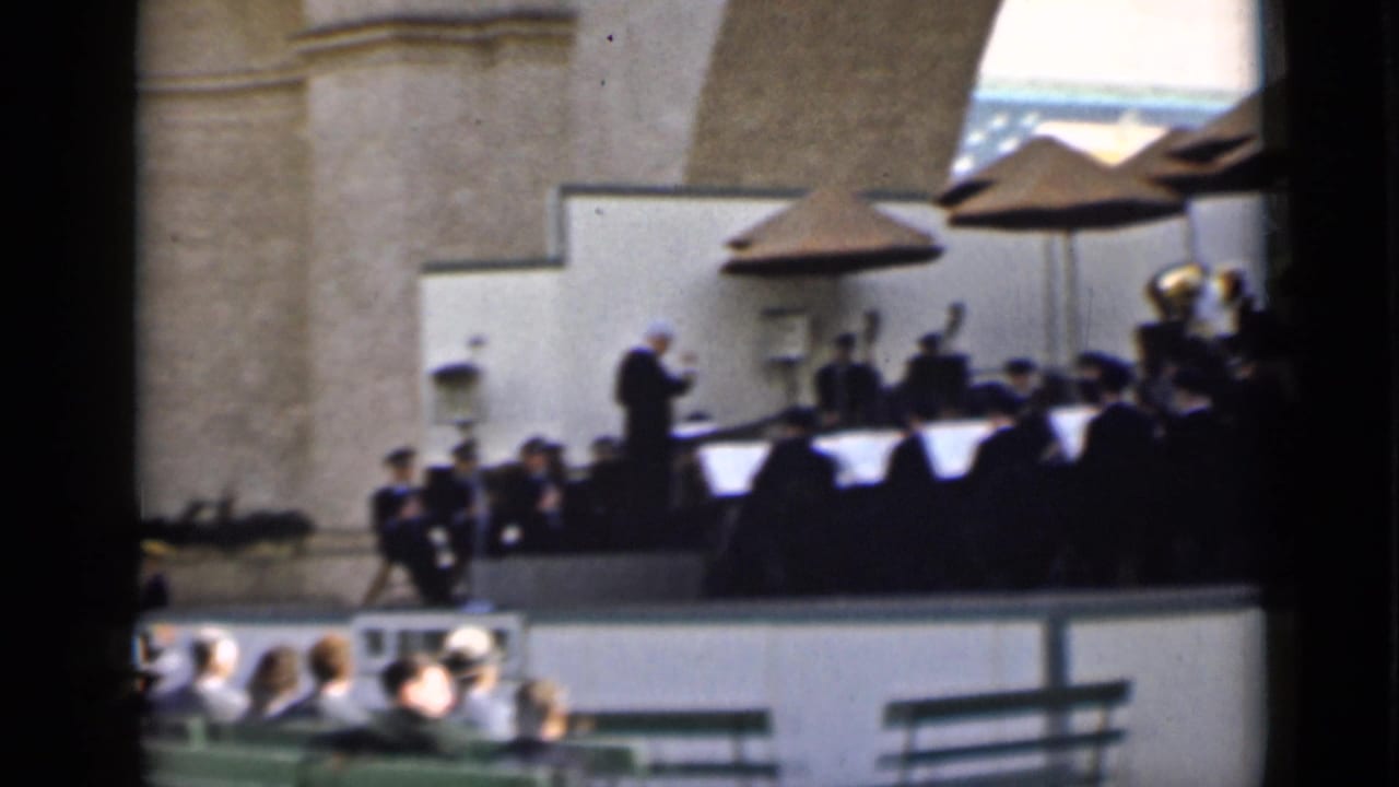 A conductor stands before musicians on a raised outdoor stage in San Francisco, with an audience seated beneath the bandstand.