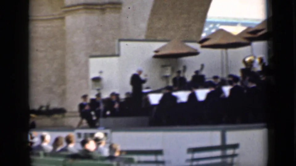 A conductor stands before musicians on a raised outdoor stage in San Francisco, with an audience seated beneath the bandstand.