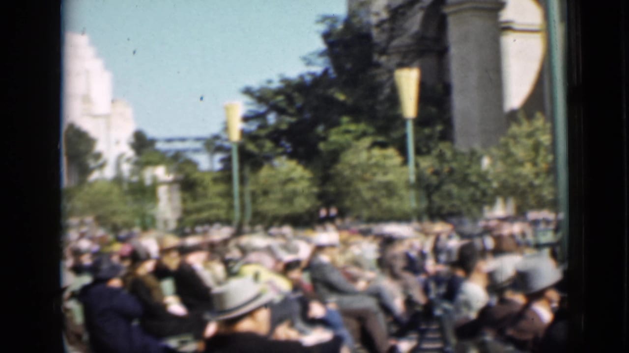 Large seated crowd in outdoor stands with trees, lampposts, and a pale building beyond.