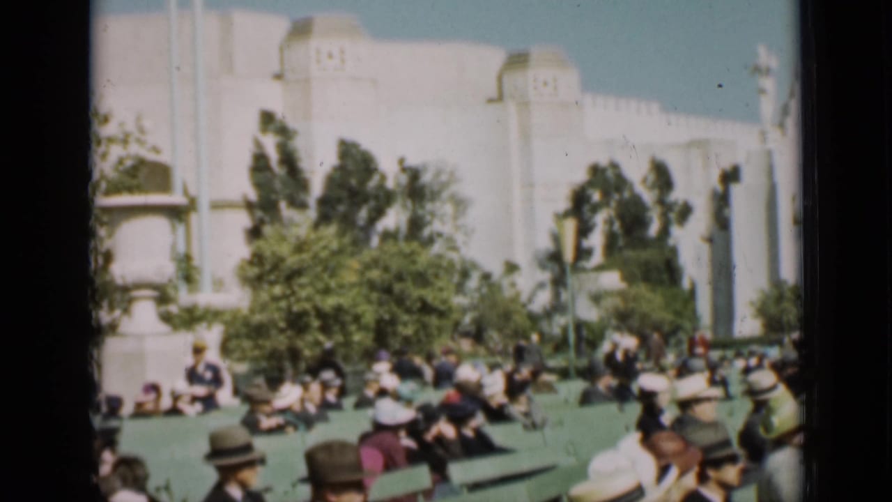 Crowd in hats seated and walking near a large white building and trees.