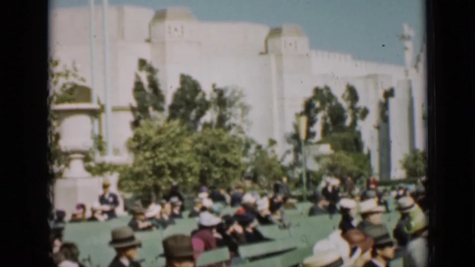 1937 San Francisco home-movie frame showing a crowd in period hats near a large white building and landscaped trees.